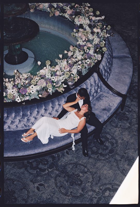Juan David leaning over Hannah on a velvet banquette beside a flower-ringed fountain, seen from above