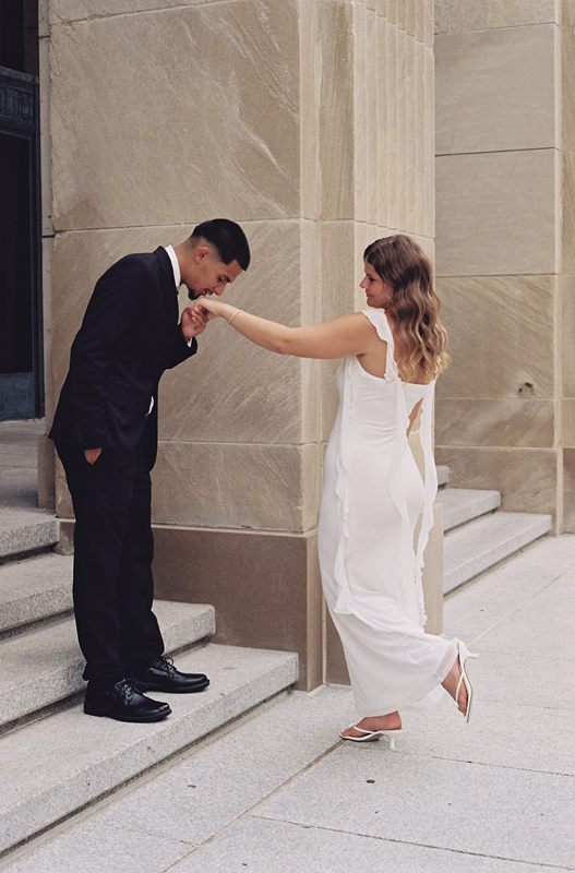 Juan David kissing Hannah's hand beside a stone column