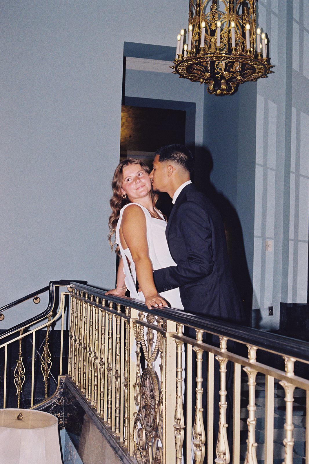 Juan David kissing Hannah's cheek at a brass railing beneath a chandelier